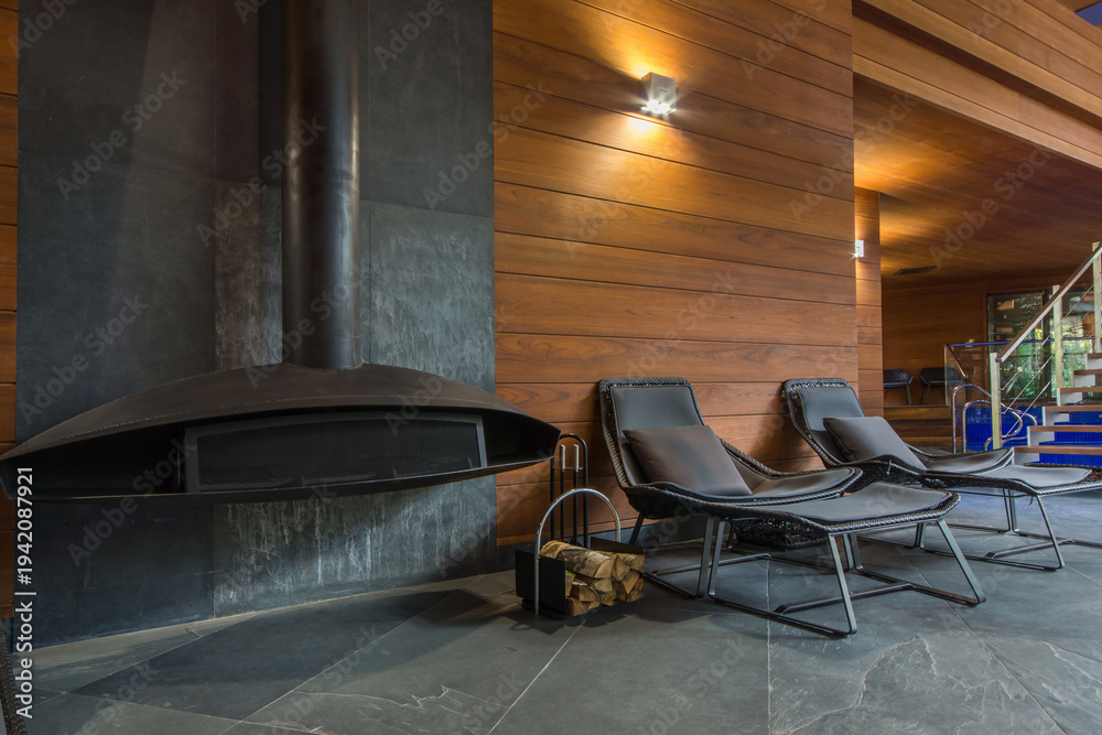 Fototapeta premium A fireplace sits on a stone wall in a space with wooden walls. Two lounge chairs are placed nearby. There are wood logs in a basket beside the fireplace. Light enters through nearby windows