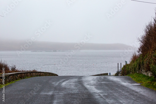 A quiet road heading into the mist at Clifden Bay, Ireland. Moody coastal scenery in Connemara with damp pavement, traditional stone walls, and a thick layer of fog over the Atlantic ocean.