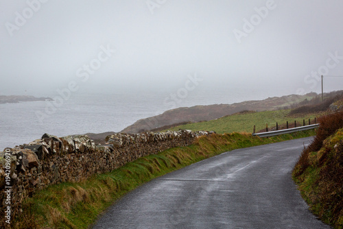 A scenic coastal road in Clifden, Ireland, curving alongside a traditional stone wall. Moody atmospheric landscape of the Connemara coastline with sea mist and rolling green hills