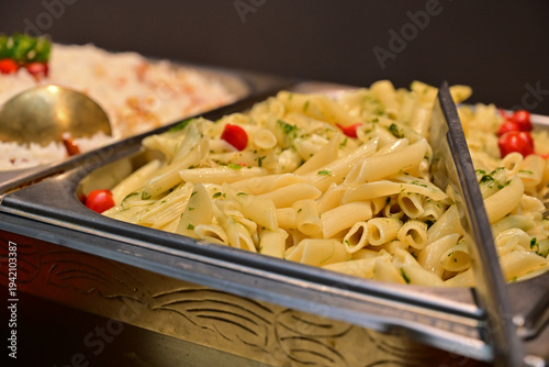 pasta, penne pasta with herbs and cherry tomatoes in buffet server, delicious italian pasta dish at catering station, gourmet pasta salad with parsley and tomatoes for event, cooked penne pasta 