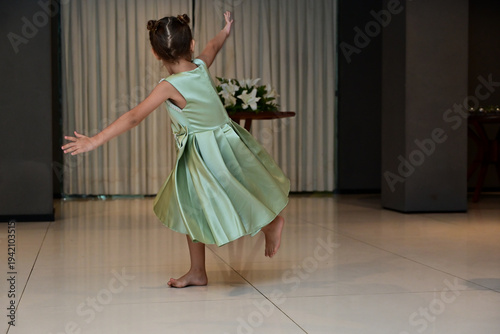 child spinning in a green dress on a tiled floor, back view of girl twirling barefoot at a formal event, little girl with open arms dancing indoors with flowers in the background, young dancer 