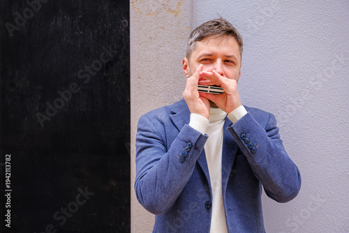 Man musician in a blue blazer and white sweater playing blues on a harmonica with eyes closed, standing outdoors in city street