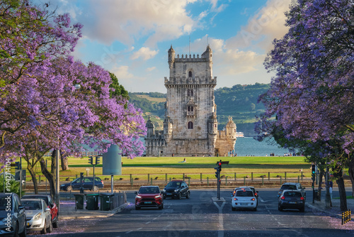 Scenic view of Belem Tower in Lisbon highlighted by blooming jacaranda trees and sailboats on the Tagus River during sunset. Belem, Lisbon, Portugal