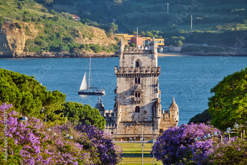 Belem Tower stands majestically above blooming jacaranda trees with tourists sailing on the Tagus River at sunset in Lisbon.