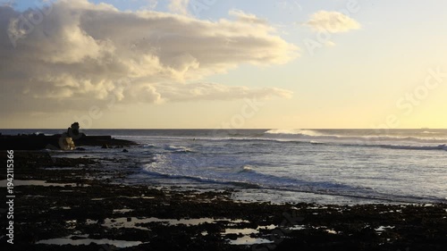 Golden hour waves rolling onto the rocky shoreline on Tenerife, with warm sunset light illuminating the Atlantic coast and a silhouetted rock formation along the water’s edge