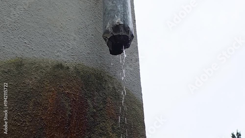 Deteriorated downspout spilling steady droplets onto surface. Industrial style scene showing water leaking from aging pipe onto gritty concrete with rust marks and wet stones