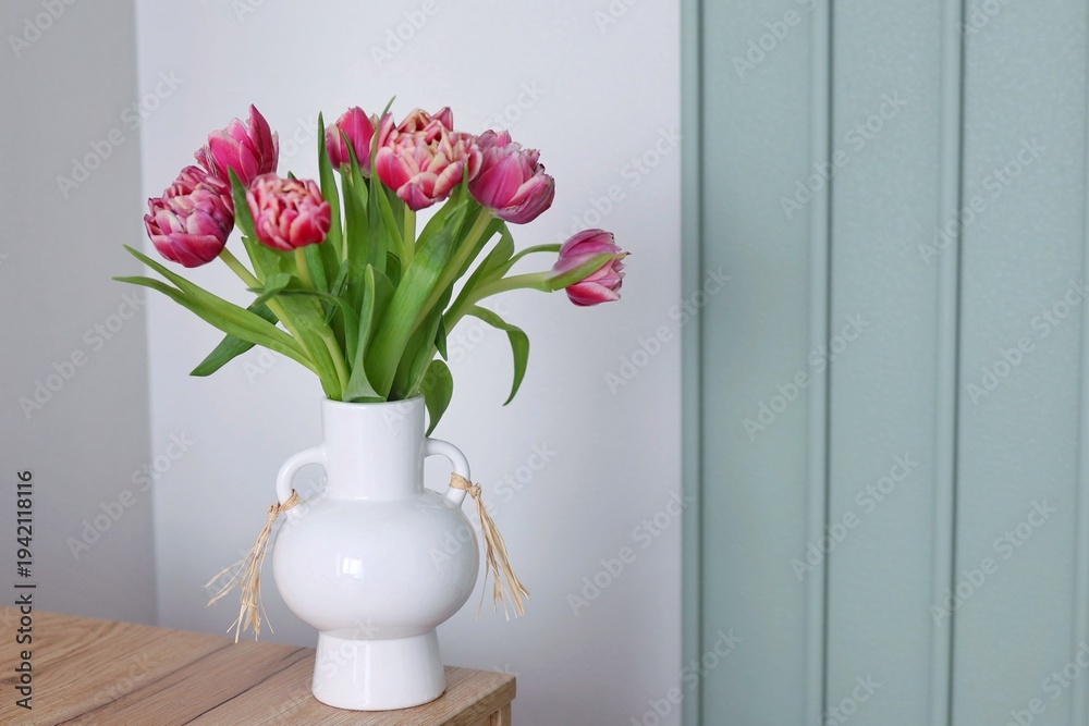Fototapeta premium A beautiful bouquet of pink double tulips in a white ceramic vase stands on a wooden table against a sage green paneled wall. Concept of modern spring home decor.