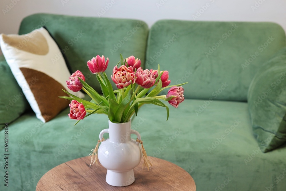Fototapeta premium A bouquet of pink double tulips in a white ceramic vase on a wooden table in front of a green velvet sofa. Concept of cozy spring home decor. 