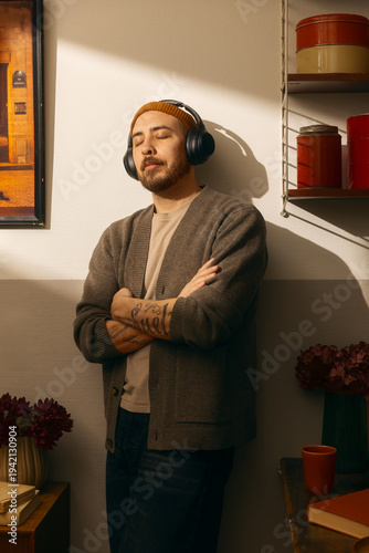 Relaxed man with beard and tattoos wearing headphones and beanie, standing with eyes closed in a cozy room with warm sunlight, leaning against a wall while listening to music or a podcast at home.