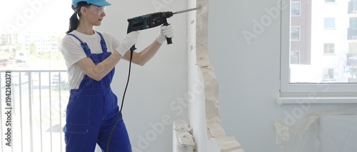 Female construction worker demolishing interior wall with rotary hammer, wearing safety glasses and hardhat, creating substantial concrete debris pile during demolition process