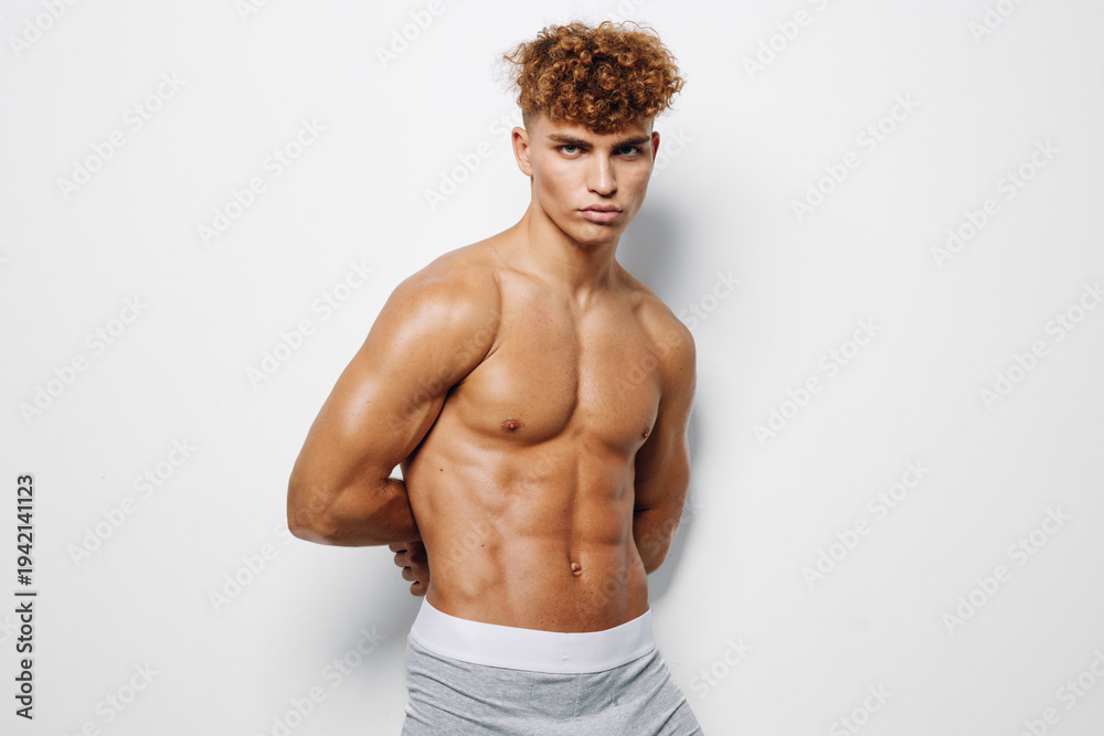 Fototapeta premium Muscular young man with curly hair posing shirtless against white background, showing defined abs and toned upper body in fitness and health concept.