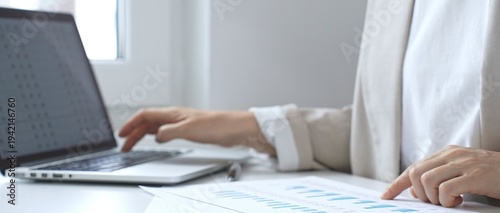 Professional female analyzing financial reports, reviewing charts on laptop and paper documents while seating at the desk in office. Business people
