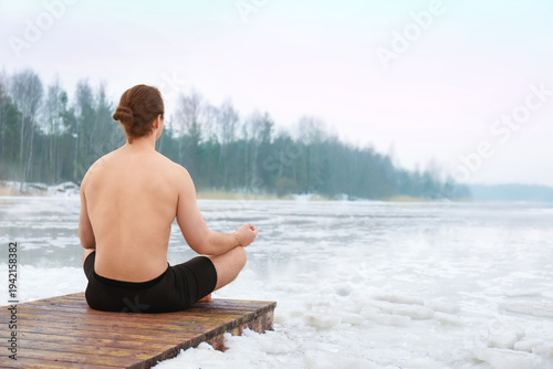 Photos Shirtless man with long hair in a bun sitting in a lotus meditation pose on a wooden pier by a frozen, icy lake during winter