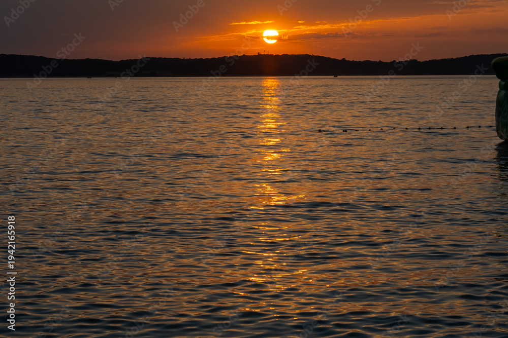 Obraz premium Intense fiery sunset with golden sun path reflecting on dark rippling sea surface and hill silhouette in deep orange sky. Concept of romantic evening mood, mystery and raw natural power.