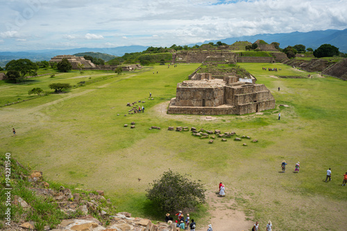 Tourists explore ancient ruins under cloudy sky in Oaxaca, Mexico, visiting Monte Alban's stone structures surrounded by green grass and distant hills.