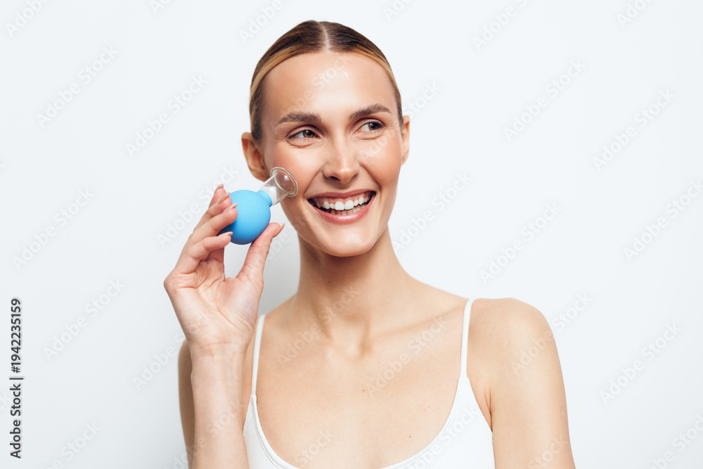 Naklejka premium Smiling woman with clear skin using a facial cleansing brush on white background. Concept of skincare, beauty, personal care, and healthy lifestyle. Bright studio lighting highlights natural look.