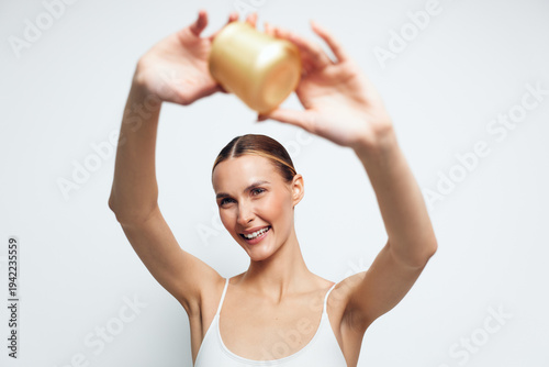 Foto A happy young woman with slicked back hair holds a jar of cream above her head smiling in a bright studio