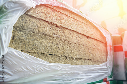 Stack of mineral wool thermal insulation material wrapped in protective plastic film at a construction site or warehouse.
