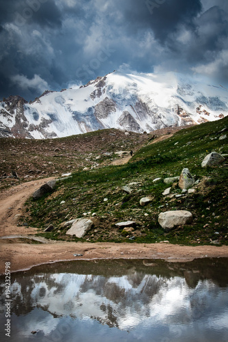 Scenic reflection of Molodezhny Glacier in a mountain puddle. Dramatic clouds over snow-capped peaks in Trans-Ili Alatau. Artistic alpine nature photography.