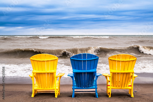 Canvas Print Yellow and blue Adirondack chairs on a sandy Toronto beach facing crashing waves of Lake Ontario during a windy spring day