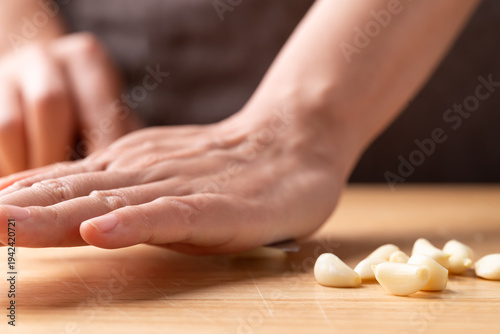 Fotografía Hand crushing fresh garlic clove with a knife on a wooden cutting board, prepare