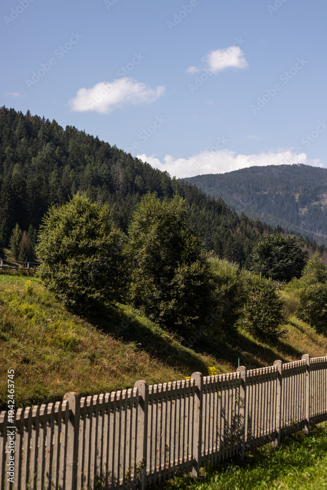 Fototapeta premium A peaceful rural scene featuring a rustic wooden fence along a grassy slope, with lush green trees and rolling forested mountains under a bright blue sky with scattered clouds.