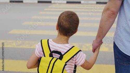 School run with father, Little boy’s morning routine, Dad ensuring child safety, Urban crosswalk with family, Yellow backpack for visibility, Responsible parenting in action, Together on the way to