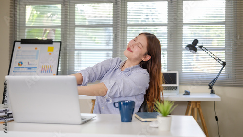 Young businesswoman stretching her arms while sitting at a desk with a laptop, taking a short break during work in a home office.