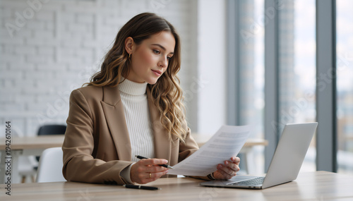 Professional woman reading document at office desk. Businesswoman reviewing paperwork with laptop in modern workspace. Corporate manager analyzing report