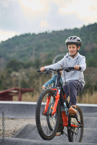 Older brother riding a mountain bike over rollers on an outdoor pump track while his younger sibling follows behind