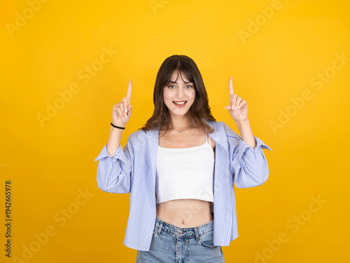 Cheerful young woman pointing fingers up at copy space, looking at camera with happy smile, isolated on yellow background. Female showing blank space for text, logo or advertisement, promotion concept