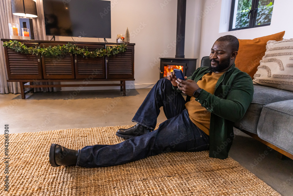 Obraz premium African American adult man lounging on rug in living room in sweater using smartphone, copy space