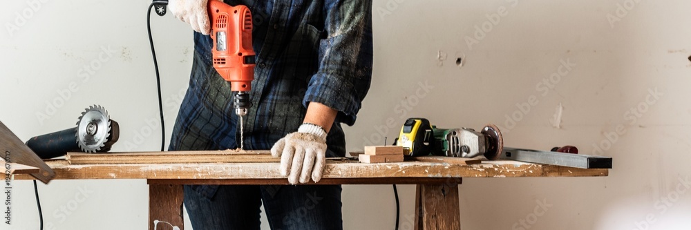 Obraz premium Person using a drill on a wooden plank. Tools on a workbench. DIY project with drill, wood, and tools. Hands-on work with drill and wood. Construction scene. Home construction and renovation concept.