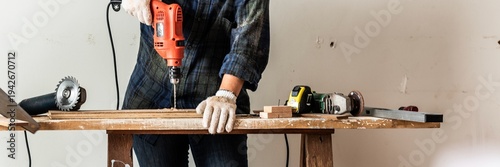 Person using a drill on a wooden plank. Tools on a workbench. DIY project with drill, wood, and tools. Hands-on work with drill and wood. Construction scene. Home construction and renovation concept.