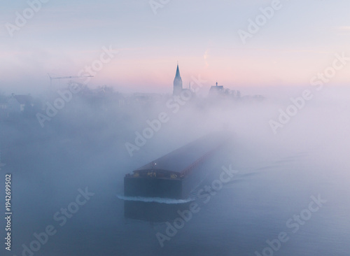Aerial view of a misty river scene with a barge sailing through, the spire and buildings barely visible in the background, Beesel, Limburg, Netherlands.