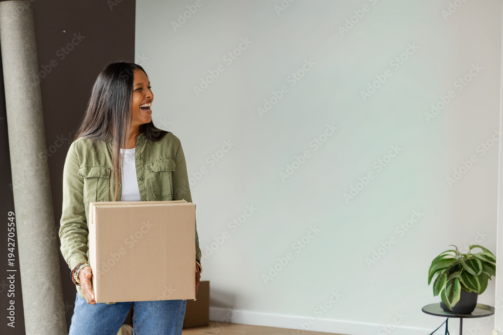 Obraz premium Mid-adult Indian woman holding cardboard box, wearing green jacket and jeans in minimal room