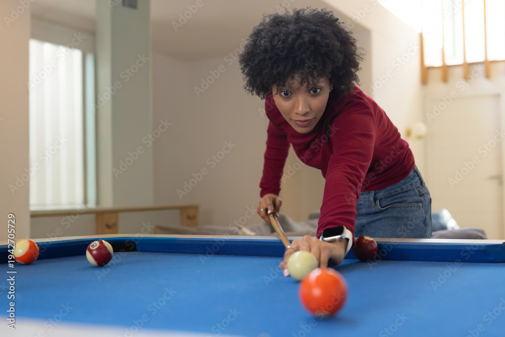 Fototapeta premium Adult African-American woman in jeans leaning over home pool table holding cue lining up cue ball