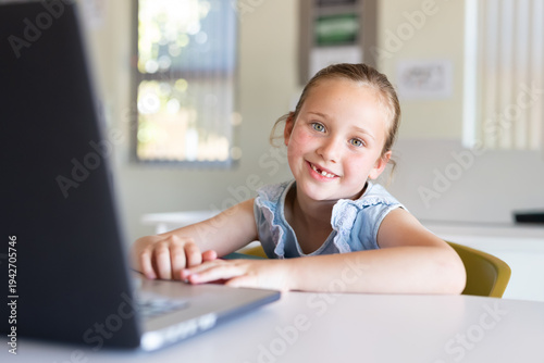 Girl sitting at table in classroom working on laptop, smiling with missing tooth, wearing blue top