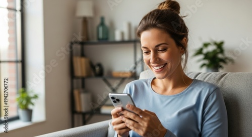Joyful Woman with Top-Knot Bun Smiling at Smartphone in a Sunlit, Cozy Living Room.