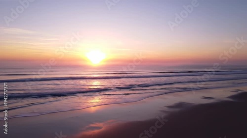 Sunrise over calm ocean waves on sandy beach, vibrant colors reflecting on water surface, creating a serene coastal atmosphere at dawn
