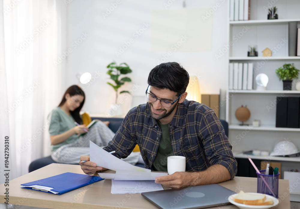 Obraz premium A young man smiles as he reviews documents on his laptop, while his partner relaxes behind him. The atmosphere is warm and inviting in this home office, reflecting the concept of working from home.