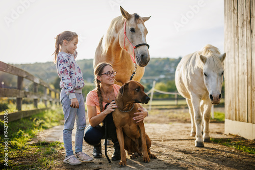 Portrait of woman and her daughter with dog and horses on family ranch at sunset. Rural lifestyle with pets and animals. Happy moment together