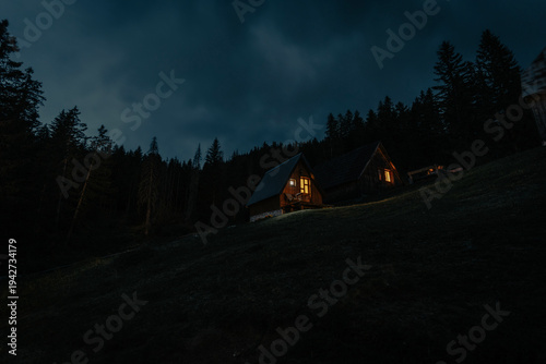 Fotografie Two cozy mountain chalets with glowing warm windows against a dark dense forest and night sky