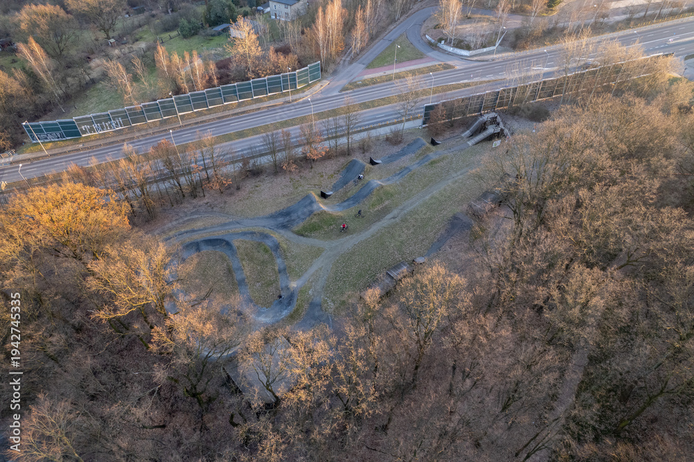 Fototapeta premium Aerial view of a bike pump track and forest recreation area next to an urban road at sunset.