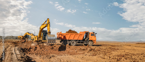 Heavy machinery is moving dirt at a construction site. A dump truck is loading soil while an excavator works nearby. The sun shines in a clear sky