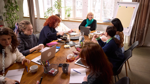 Women business team working together at large wooden table. Concept of inclusive workplace culture, leadership guidance, strategic planning session, corporate collaboration, professional development.