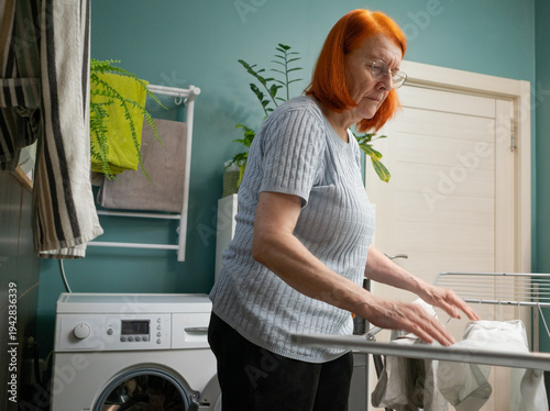 Billede på lærred From a lateral angle, a mature woman is depicted carefully hanging freshly washed items on a white drying rack during her housekeeping