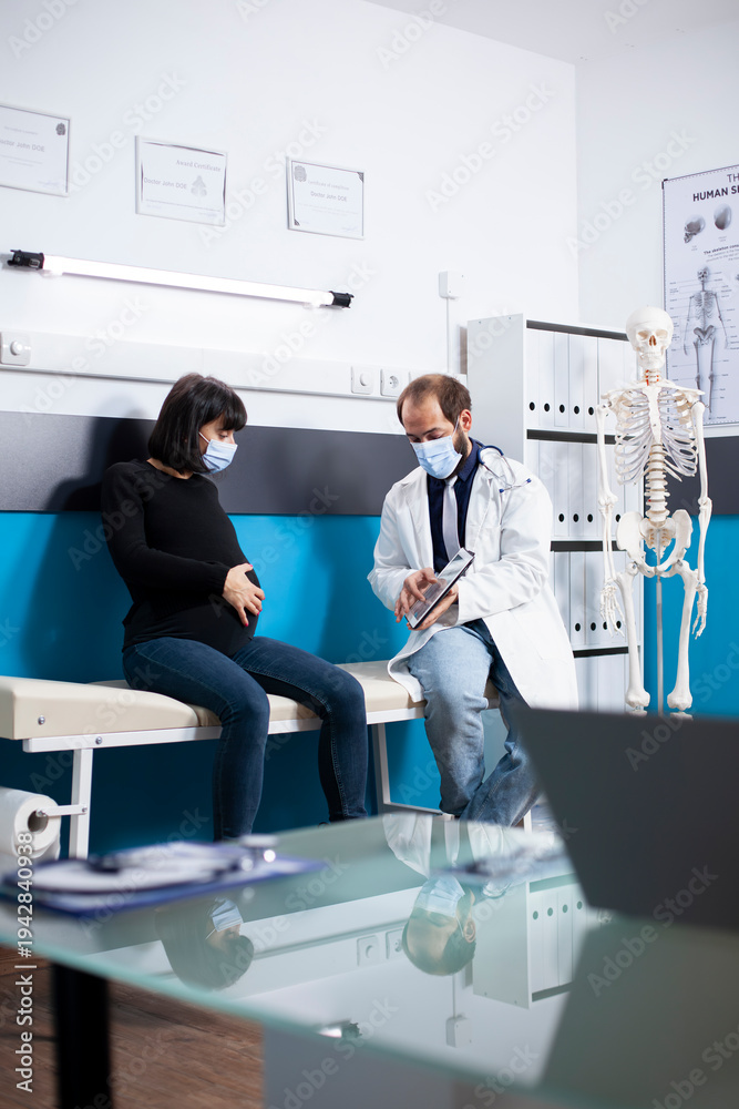Fototapeta premium In clinic, physician holds digital device to show expectant lady her medical diagnosis during pregnancy checkup. Doctor with tablet sits on hospital bed with pregnant woman, discussing maternal health