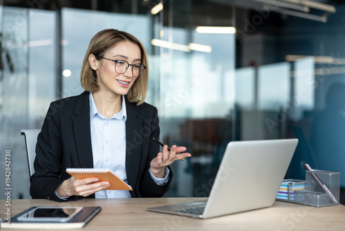 Professional businesswoman engaging in a remote video conference, taking notes in a notebook, and using a laptop in a modern office, representing virtual communication and distance learning concepts