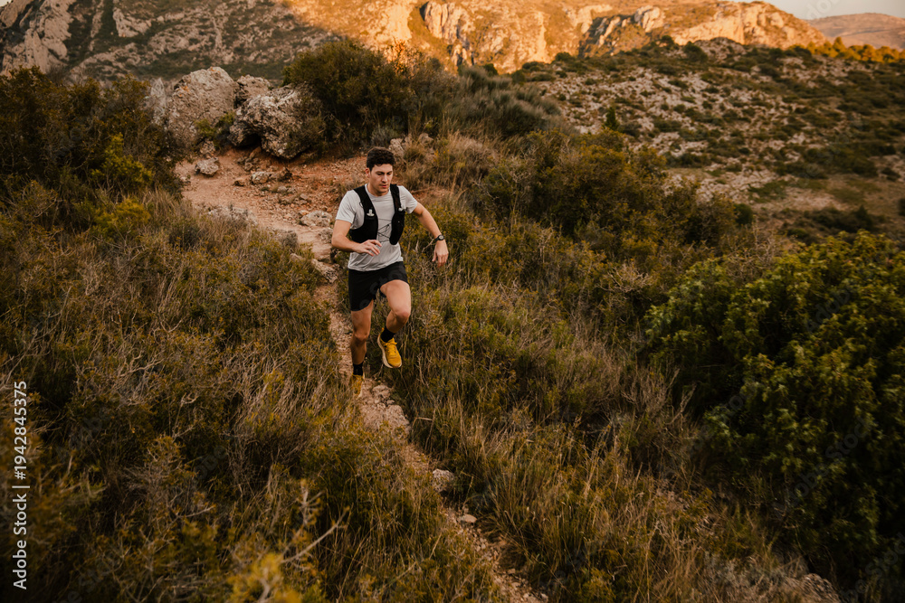 Obraz premium Athletic trail runner running along a winding mountain trail surrounded by wild vegetation and rugged terrain. Outdoor endurance sport and adventure lifestyle in nature.
